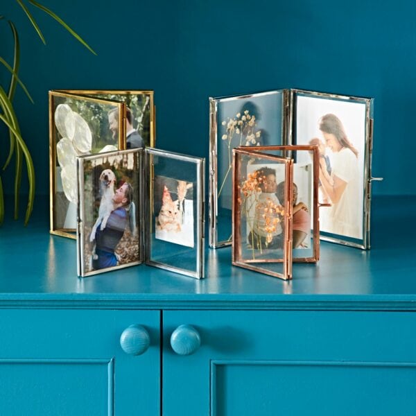 Five framed photographs arranged on a bright blue surface, featuring images of people, flowers, and a dog. The photo frames come in various sizes and finishes, with some showcasing the stunning Recycled Glass Folded Double Photo Frame design.