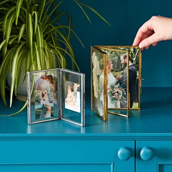 A hand adjusts a gold frame with a wedding photo on a blue cabinet. Nearby, the Recycled Glass Folded Double Photo Frame in silver holds multiple small photos. A potted plant is in the background, adding a touch of green.