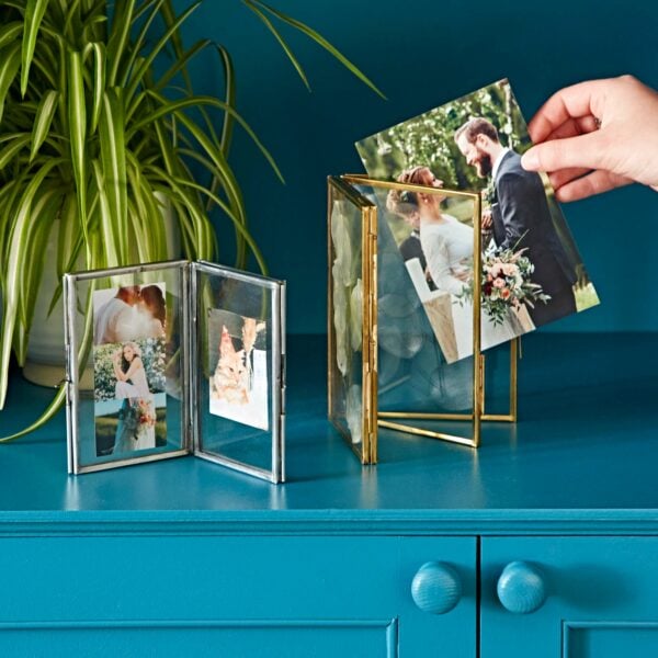 A hand places a photo of a bride and groom in a gold picture frame on a blue cabinet, beside the Recycled Glass Folded Double Photo Frame and a potted plant.