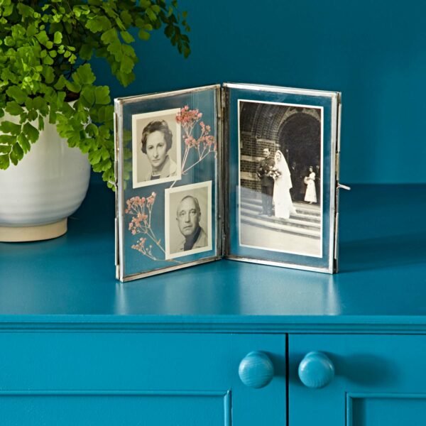 The Recycled Glass Folded Double Photo Frame, placed on a teal dresser, elegantly holds black-and-white portraits on the left and a wedding photo on the right. In the background sits a potted plant.