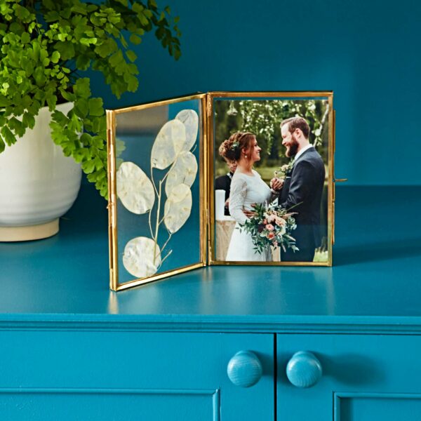 A framed photo of a couple in wedding attire holding hands is placed on a blue dresser with a potted fern in the background. The picture sits elegantly within the Recycled Glass Folded Double Photo Frame, adding an eco-friendly touch to the romantic scene.