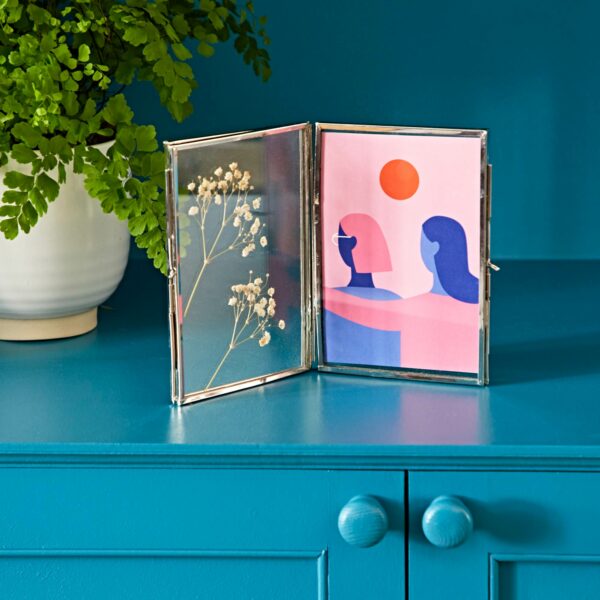 A Recycled Glass Folded Double Photo Frame on a teal cabinet displays pressed flowers on one side and an abstract illustration of two people and a sun on the other, with a potted plant in the background.