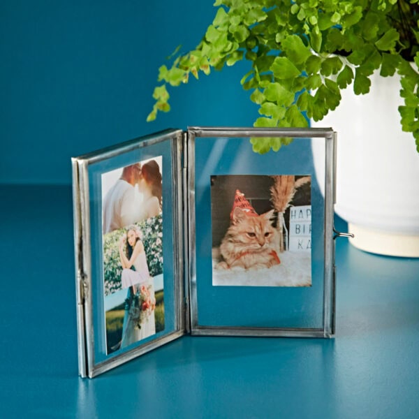 The Recycled Glass Folded Double Photo Frame can elegantly display two cherished photos: a couple sharing a kiss on the left, and a playful cat in a party hat on the right. In the background, you can see a leafy plant in a white pot, adding a touch of nature to the scene.