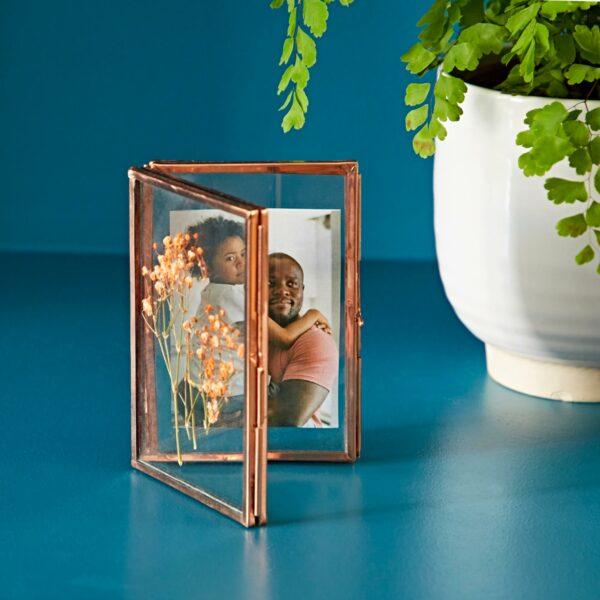 A Recycled Glass Folded Double Photo Frame, showcasing a dried flower and a photo of a man and a child, is placed on a blue surface next to a potted fern.