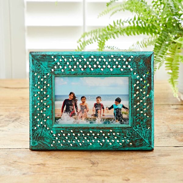 A 6" x 4" Hara Green Distressed Mango Wood Photo Frame beautifully displays a photo of four children enjoying the ocean waves, with intricate decorative patterns adding charm and a potted fern in the background bringing a hint of nature.