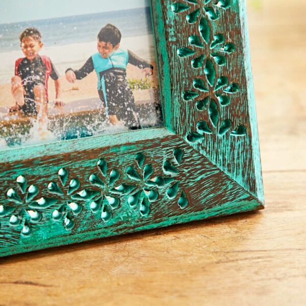 The 6" x 4" Hara Green Distressed Mango Wood Photo Frame, featuring turquoise paint and intricate floral carvings, holds a picture of two children playing near water. It is placed on a wooden surface.