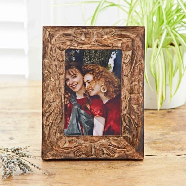 The 6" x 4" Jaraja Natural Mango Wood Photo Frame elegantly displays a picture of two smiling women with long hair—one with straight hair and the other with curly hair. The scene is complemented by a potted plant and dried flowers in the background, enhancing its natural charm.