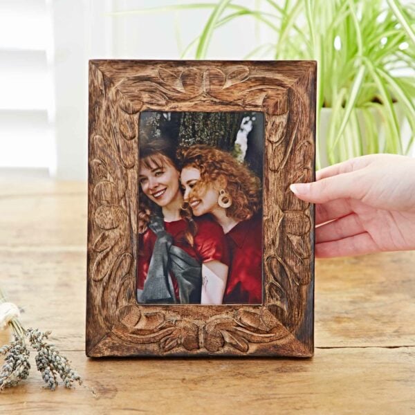 A hand is holding the 6" x 4" Jaraja Natural Mango Wood Photo Frame, showcasing a picture of two smiling people, one with red curly hair, standing close together. A green plant provides a vibrant background. This stunning photo frame made of natural mango wood adds a touch of warmth to any room.