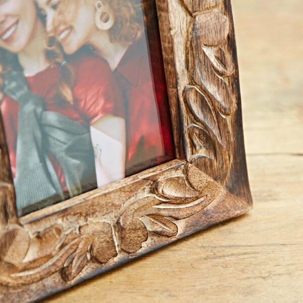 A 6" x 4" Jaraja Natural Mango Wood Photo Frame, adorned with intricate carvings, sits on a wooden table, containing a photograph of two women smiling and standing close together.
