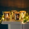 Four lit candles in a set of silver antique effect tea light holders sit on a white surface, surrounded by illuminated green leaves, creating a warm, decorative scene.