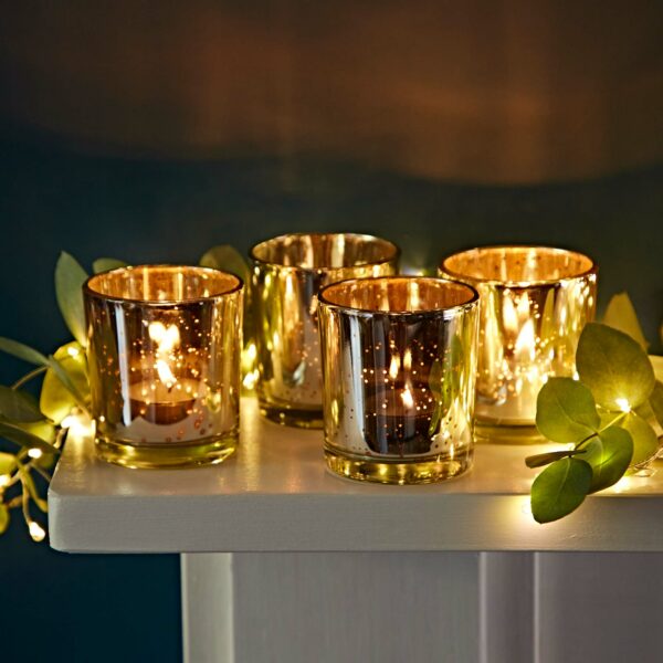 Four lit candles in a set of silver antique effect tea light holders sit on a white surface, surrounded by illuminated green leaves, creating a warm, decorative scene.