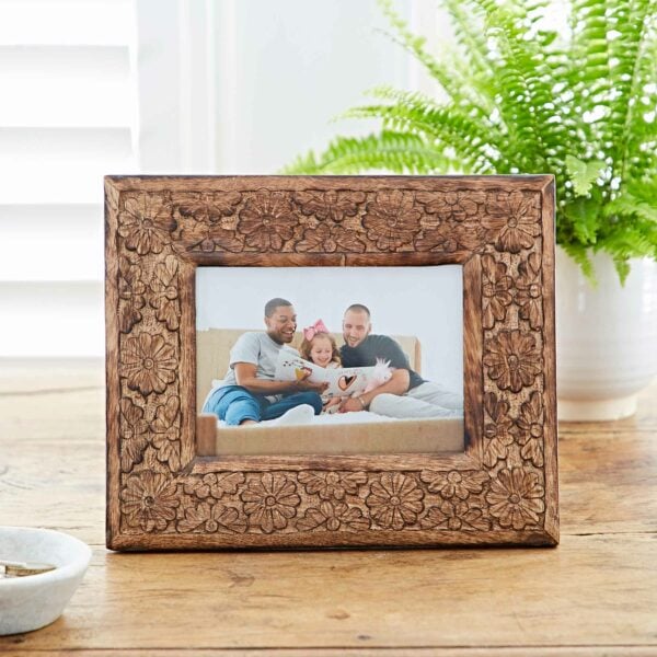 The 7" x 5" Asali Natural Floral Pattern Mango Wood Frame holds a photo of three people sitting on a couch, smiling and holding a child. In the background, there is a potted fern on a wooden table.