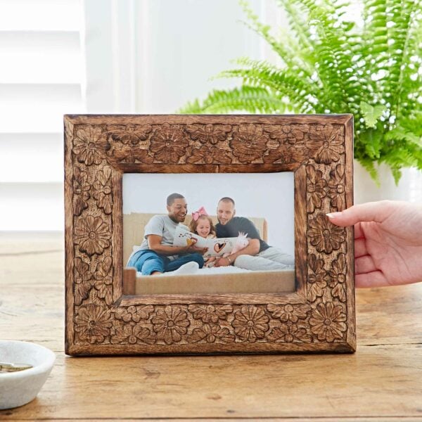 A 7" x 5" Asali Natural Floral Pattern Mango Wood Frame displays a picture of three smiling adults and a child sitting on a couch. The frame is being held up by a hand, with a plant in the background.