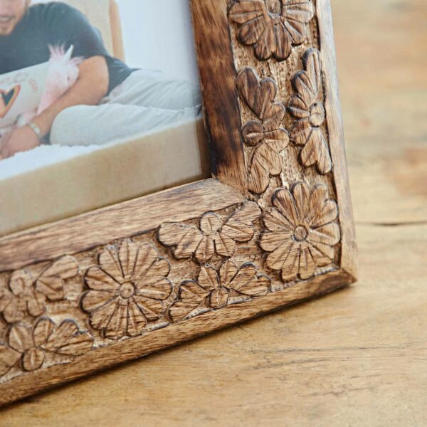 Close-up of the 7" x 5" Asali Natural Floral Pattern Mango Wood Frame with intricate carved floral and butterfly designs, set on a wooden surface. The edge of a photograph within the frame is partially visible.