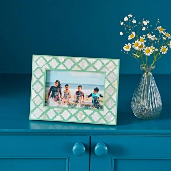 A Kajol Mother of Pearl 6" x 4" photo frame holding a beach photo of four children sits on a blue cabinet beside a glass vase filled with white and yellow flowers.