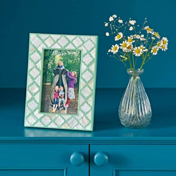 A Kajol Mother of Pearl 6" x 4" Photo Frame with a photo of a woman and three children sits on a blue cabinet next to a glass vase filled with white daisies.