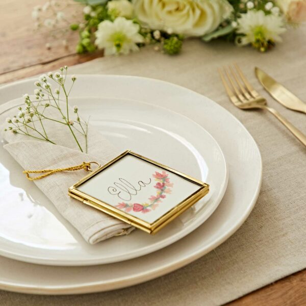 A table setting featuring white plates, a cloth napkin, silverware, and a small frame decorated with floral designs and bearing the name "Ella" placed on top of the napkin. Adjacent to this arrangement are Alia Square Glass Hanging Photo Frames adding an elegant touch.