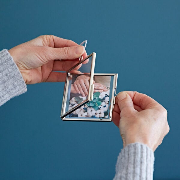 Hands holding a small framed photograph of two people holding hands, encased in one of those Alia Square Glass Hanging Photo Frames, with a blue background.
