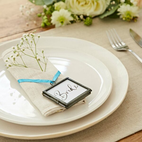 A plate setting with a napkin tied with a blue ribbon, featuring a "Bride" tag and decorated with small flowers, placed on a linen mat. Alia Square Glass Hanging Photo Frames add elegance to the scene, while white flowers create a perfect backdrop.