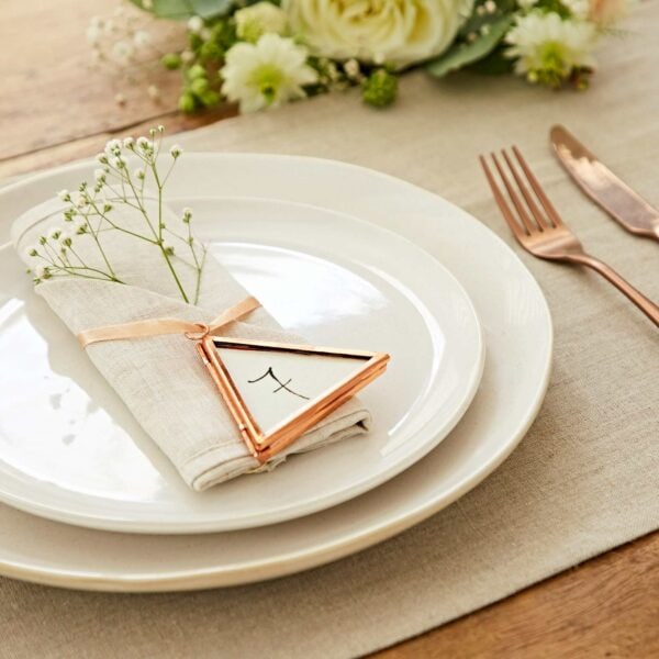 A white plate with a folded beige napkin, adorned with a small white flower in an Alia Mini Triangular Glass Hanging Photo Frame, is set on a linen tablecloth beside a rose gold fork and knife. White and green flowers are visible.
