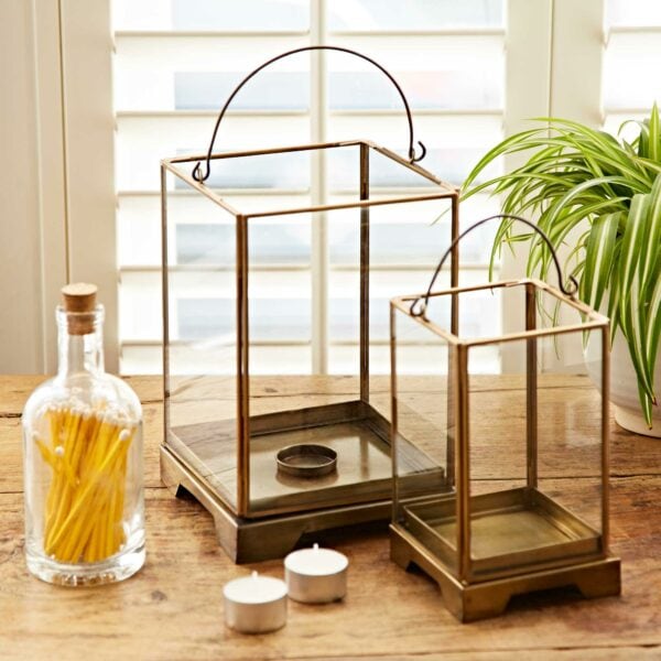 Two Antique Brass Candle Lanterns sit on a wooden table, along with a jar of matches, two tea light candles, and a potted plant.