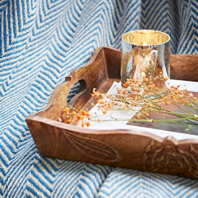 The Natural Mango Wood Tea Tray features a lit silver candle and dried flowers, set against a backdrop of blue and white patterned fabric.