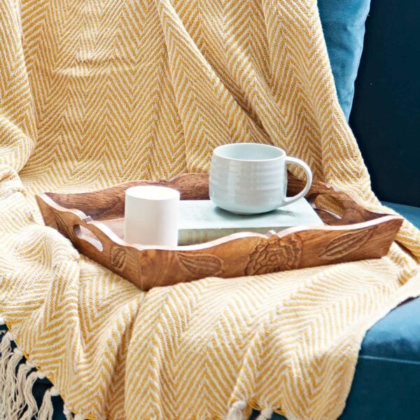 A Natural Mango Wood Tea Tray holds a mug, a small candle, and a book on a yellow herringbone-patterned blanket draped over the couch.