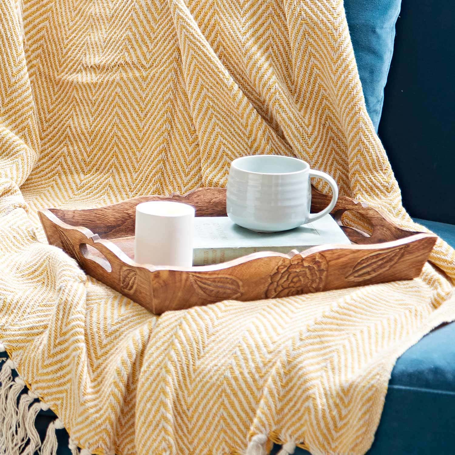 A Natural Mango Wood Tea Tray holds a mug, a small candle, and a book on a yellow herringbone-patterned blanket draped over the couch.