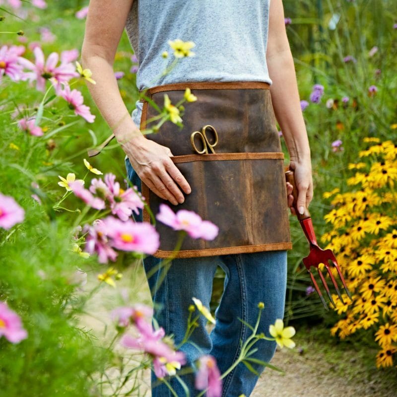 Wearing the Personalised Buffalo Leather Gardening Tool Belt, someone stands among flowers, holding a red hand fork with scissors tucked into the apron pocket.