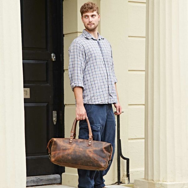 A man in a plaid shirt and dark pants stands outside a building, holding a Personalised Brown Buffalo Leather Overnight Bag by his side.
