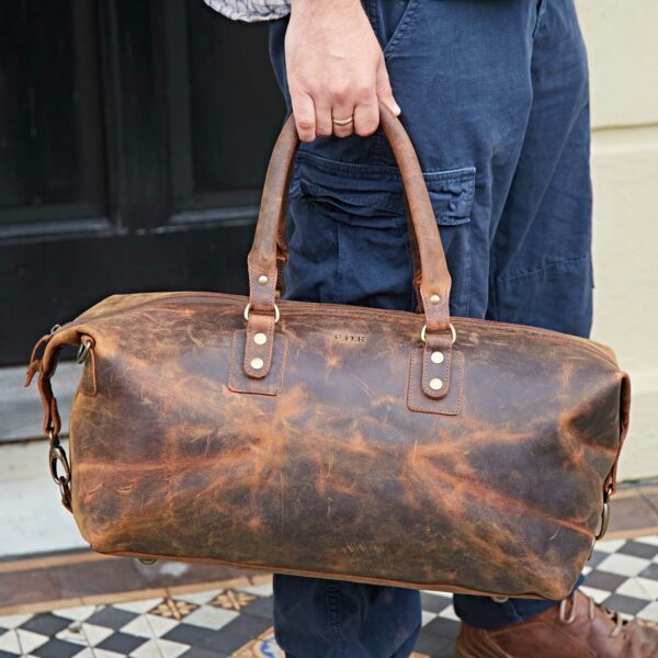 A person in blue cargo pants stands on patterned tiles, holding the Personalised Brown Buffalo Leather Overnight Bag by its handles.
