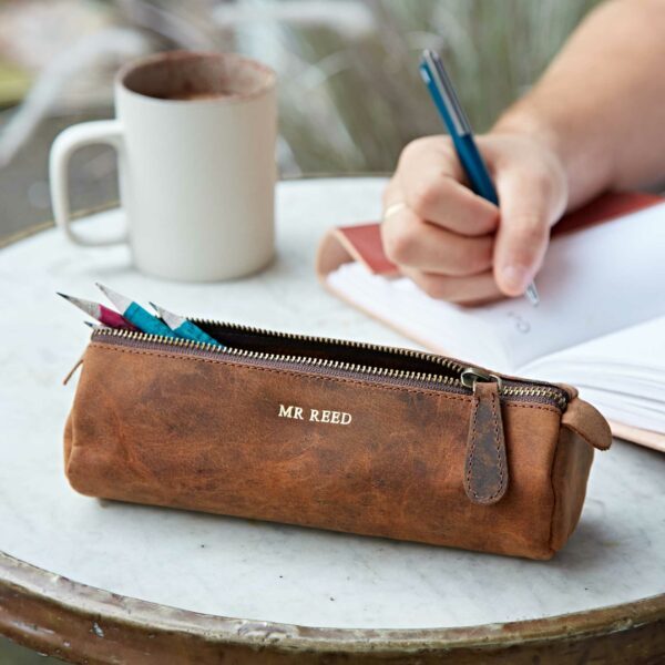A Personalised Buffalo Leather Round Pencil Case labeled "MR REED" rests on a round table next to a notebook, a mug, and a hand holding a blue pen.