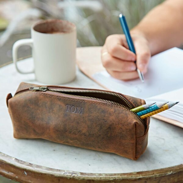 A brown buffalo leather pencil case, personalised with "TOM" embossed on it, sits on a table beside a white mug; a person writes on paper in the background.