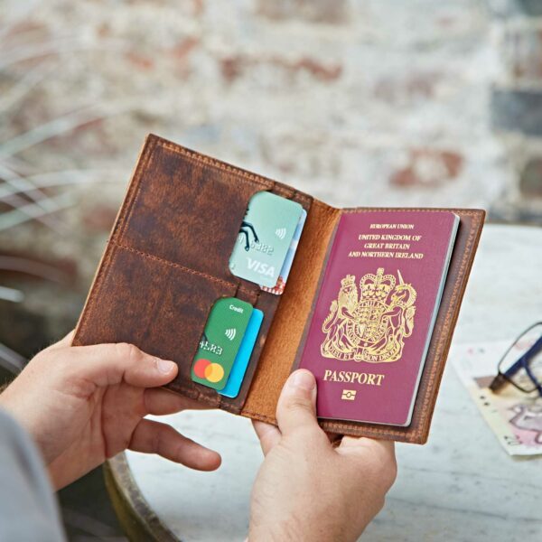 A person holds an open Personalised Buffalo Leather Passport Holder containing a UK passport and three bank cards.