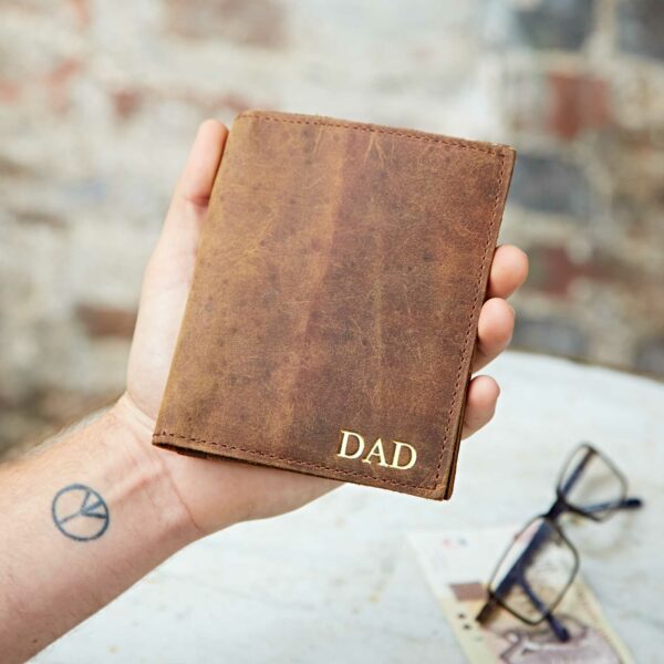 A hand holds a brown buffalo leather wallet embossed with "DAD" in gold letters; spectacles and paper rest on the table in the background, adding a touch of elegance to this personalised accessory.