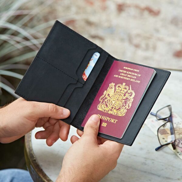 A person holds an open Black Passport Holder containing a UK passport and a card, next to a table with spectacles and a plant in the background.