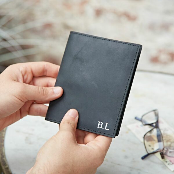 A person holds a Personalised Buffalo Leather Passport Holder, featuring the initials "B.L" embossed in white. Glasses and currency notes lie on the table nearby.