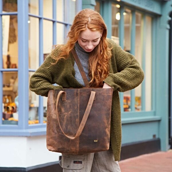 A person with long red hair looks into a Personalised Brown Buffalo Leather Tote Shopping Bag while standing on a sidewalk in front of shop windows.