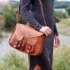 A person stands outdoors in a long-sleeved gray dress, carrying the Personalised Brown Leather Satchel Style Saddle Bag with buckle closures over their shoulder.
