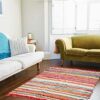 Living room with a white sofa, a green sofa, and a Multicoloured Recycled Rag Rug on the wooden floor, alongside a window adorned with white shutters.