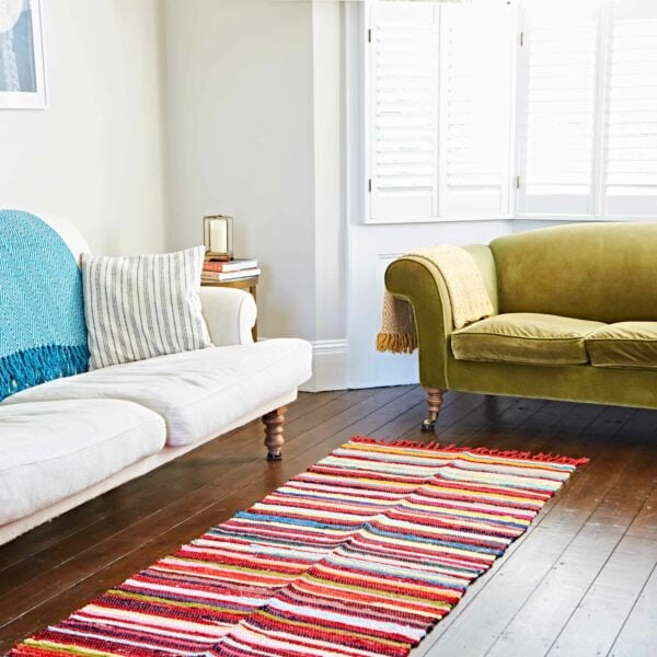 A bright living room featuring a white sofa and a green couch is adorned with a vibrant Multicoloured Recycled Rag Rug on the wooden floor, while light streams through white shuttered windows.
