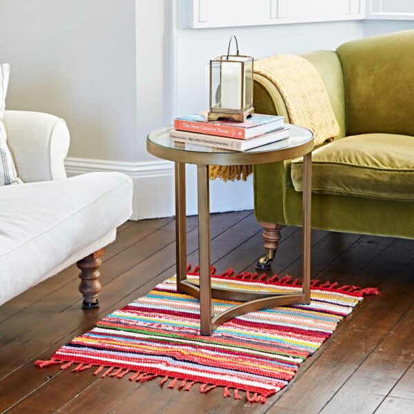 A round glass side table adorned with stacked books and a lantern sits on a vibrant Multicoloured Recycled Rag Rug, nestled between a green sofa and a white armchair in the living room.