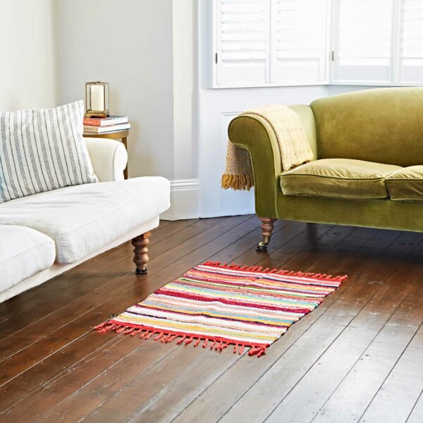 A Multicoloured Recycled Rag Rug, known for its eco-friendly design, adorns the wooden floor between a white couch and a green sofa. Nearby, a stack of books and a candle rest on a side table by the window, enhancing the cozy atmosphere.