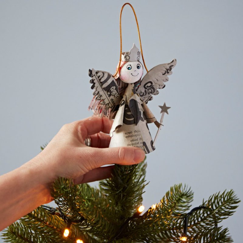 A hand places a Recycled Newspaper Angel Christmas Tree Decoration—an eco-friendly ornament made from newspaper and cardboard—on top of a Christmas tree.