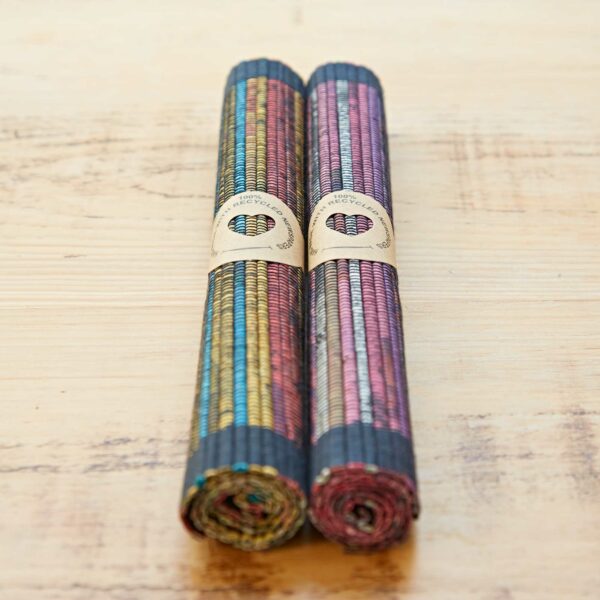 Two eco-friendly, handmade Woven Recycled Newspaper Placemats—multicolored and rolled up—lie side by side on a wooden surface, each held by a paper band.