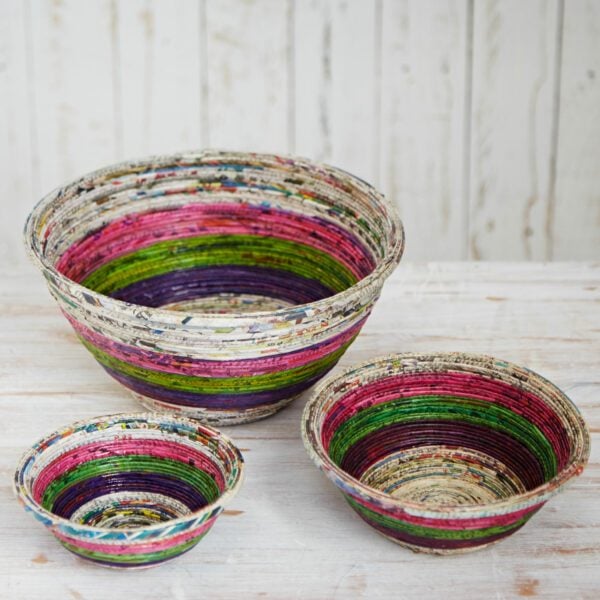 Three colourful, round bowls made from coiled, recycled newspaper are displayed on a white wooden surface, creating unique eco-friendly decor against a whitewashed wooden background.