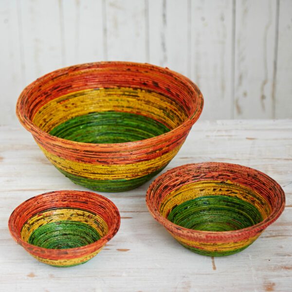 Three eco-friendly woven bowls in different sizes, featuring horizontal stripes in red, yellow, and green, are displayed on a wooden surface with a light-coloured background.