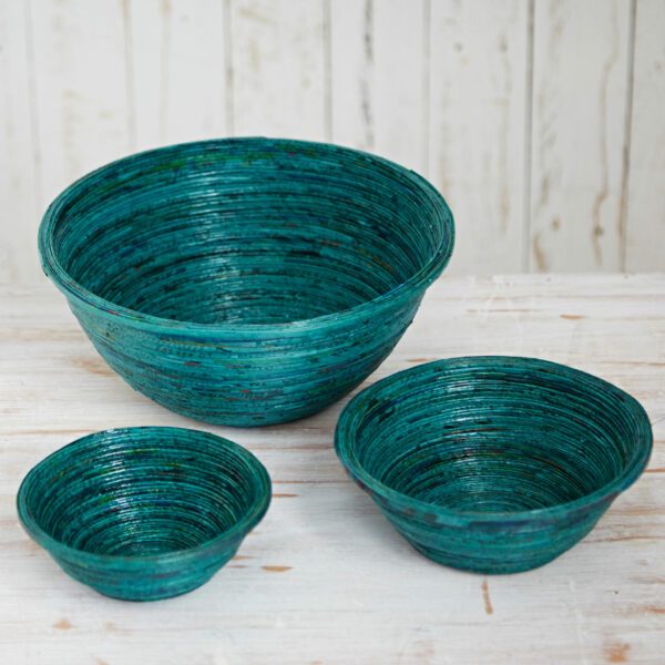 Three teal round bowls of varying sizes, crafted from recycled newspaper, are arranged on a light wooden surface with a white wooden background.