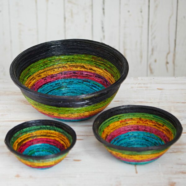 Three round bowls in different sizes, crafted from recycled newspaper, feature vibrant navy, green, yellow, pink, and blue stripes. They’re displayed on a light wooden surface against a white rustic background.