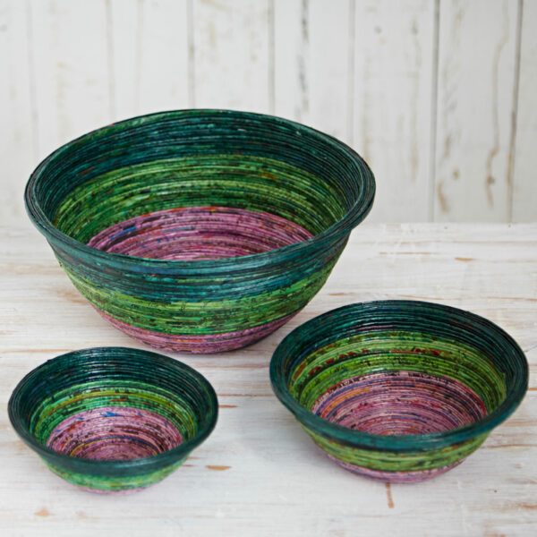 Three multicoloured round bowls, primarily green and purple, crafted from coiled material, are displayed on a light wooden surface against a rustic white backdrop—each an eco-friendly bowl made from recycled newspaper.